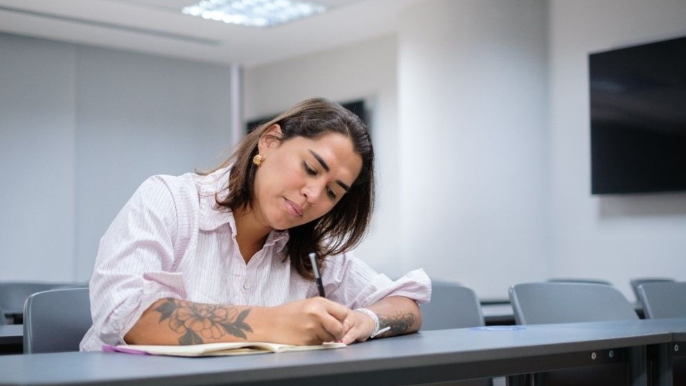 mulher com tatuagens, vestindo uma camisa branca, concentrada em escrever em um caderno em uma sala de aula moderna