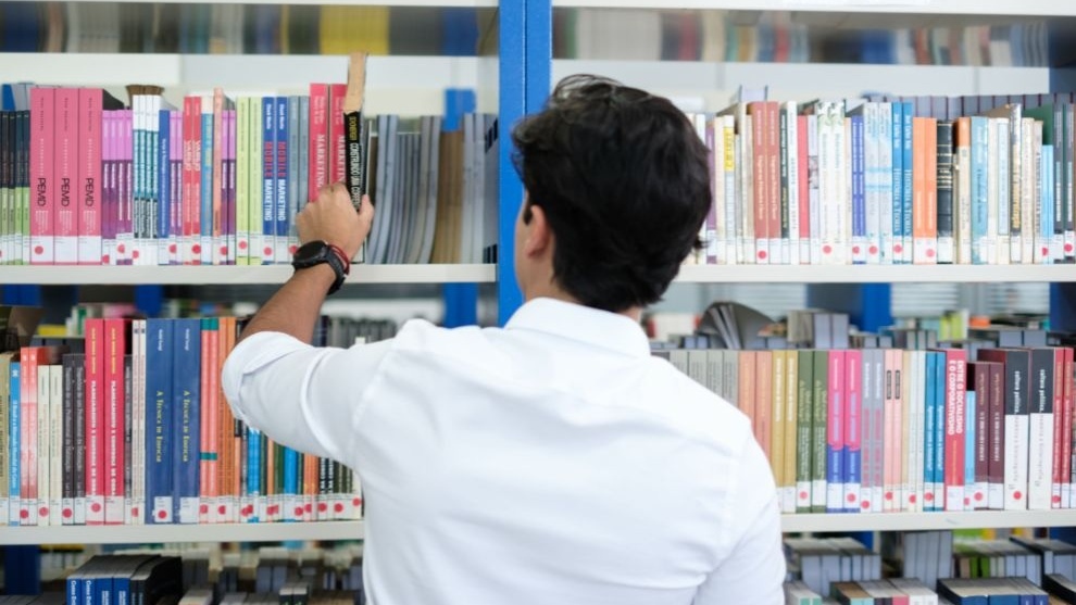 Rapaz de costas, vestindo uma camisa branca, organizando livros em uma estante de biblioteca. 