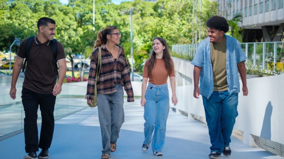 Quatro jovens caminham juntos ao ar livre em um campus, conversando e sorrindo, com árvores e prédios ao fundo.