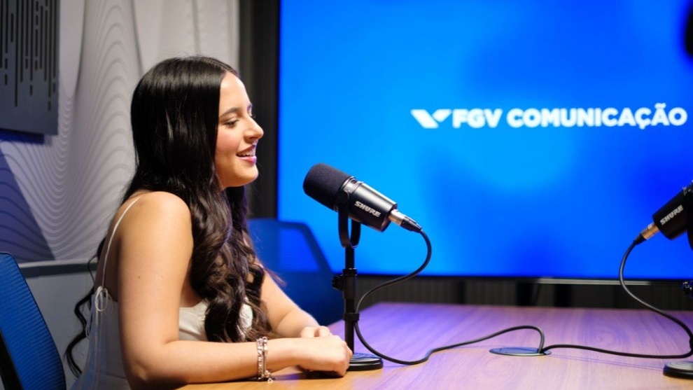 Mulher jovem falando em um microfone em estúdio de podcast, com painel azul ao fundo escrito “FGV Comunicação”.