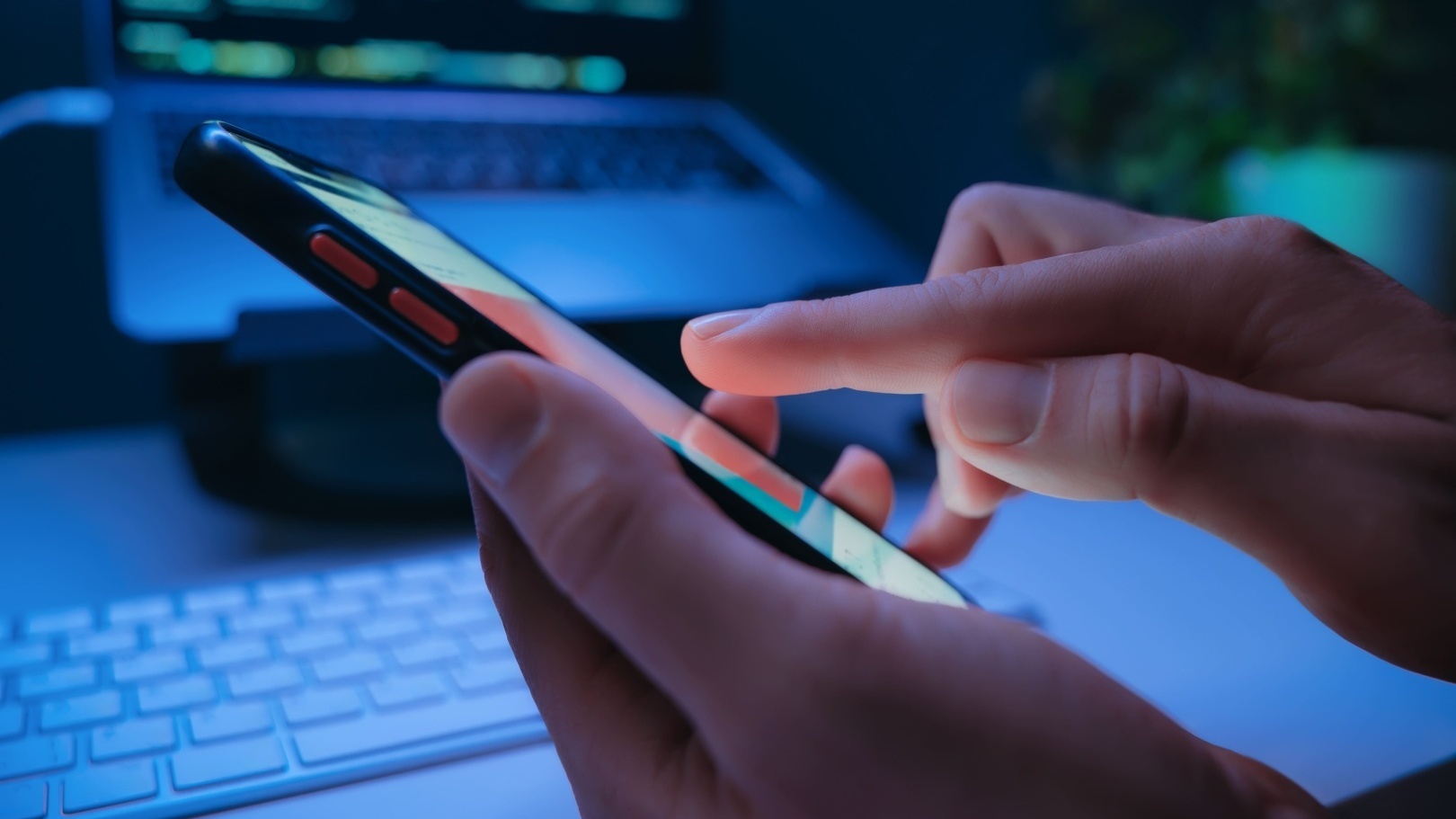 Pessoa usando um smartphone com a mão, com um notebook ao fundo exibindo telas digitais em ambiente com iluminação azul.