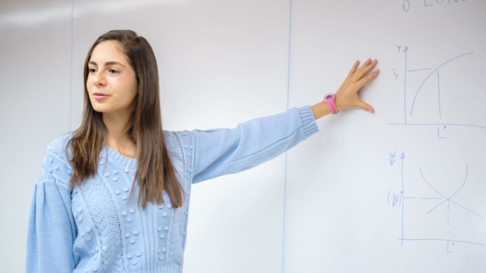 Jovem em sala de aula aponta para gráficos desenhados em uma lousa branca durante uma explicação.