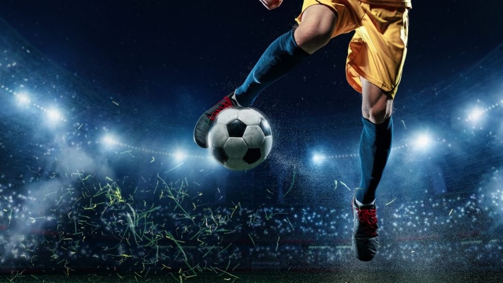 Jogador de futebol em uniforme amarelo chuta uma bola em campo iluminado por refletores de estádio.