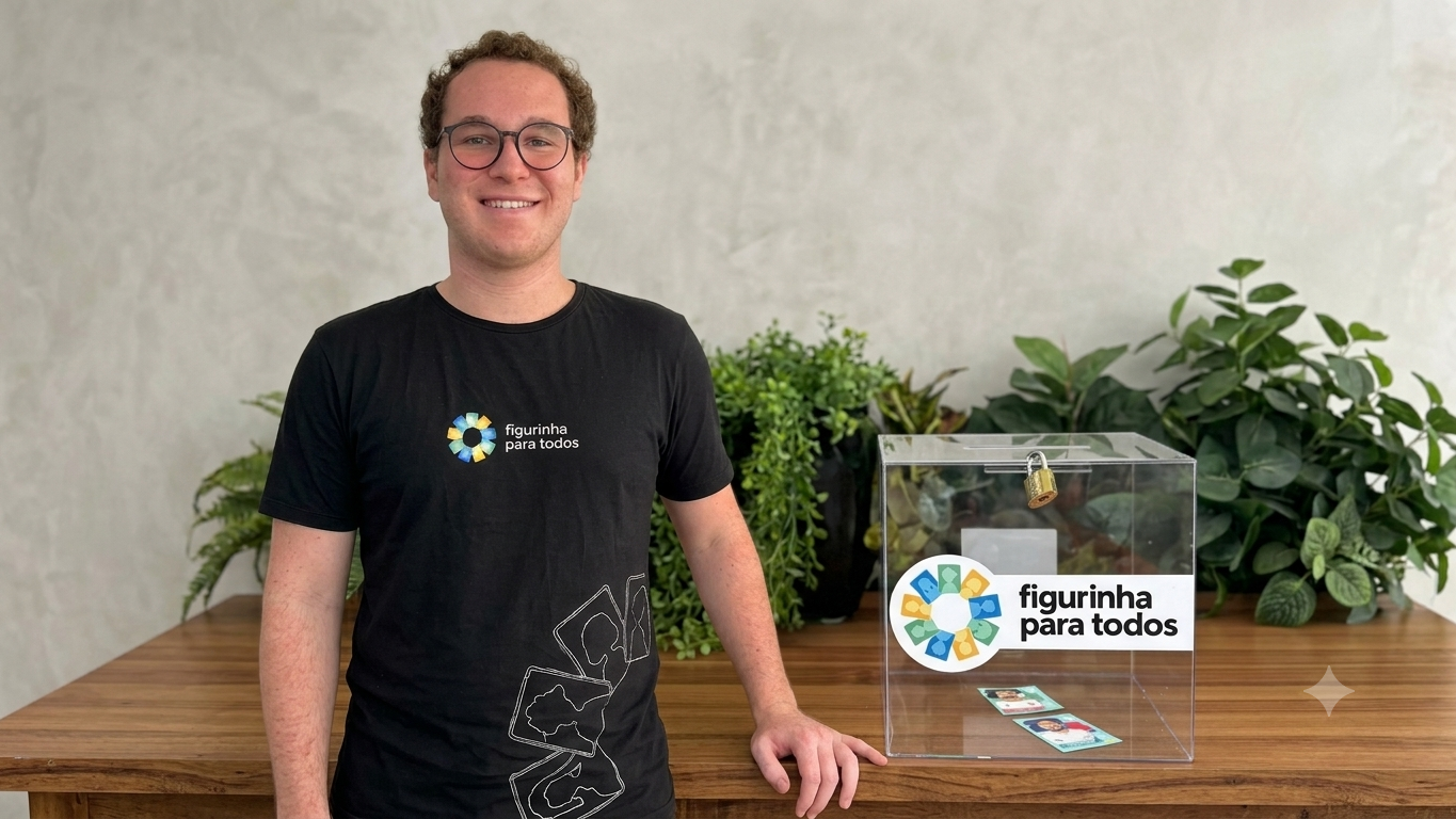 Homem sorri ao lado de uma caixa transparente com adesivo “Figurinha para Todos”, sobre uma mesa de madeira com plantas ao fundo.