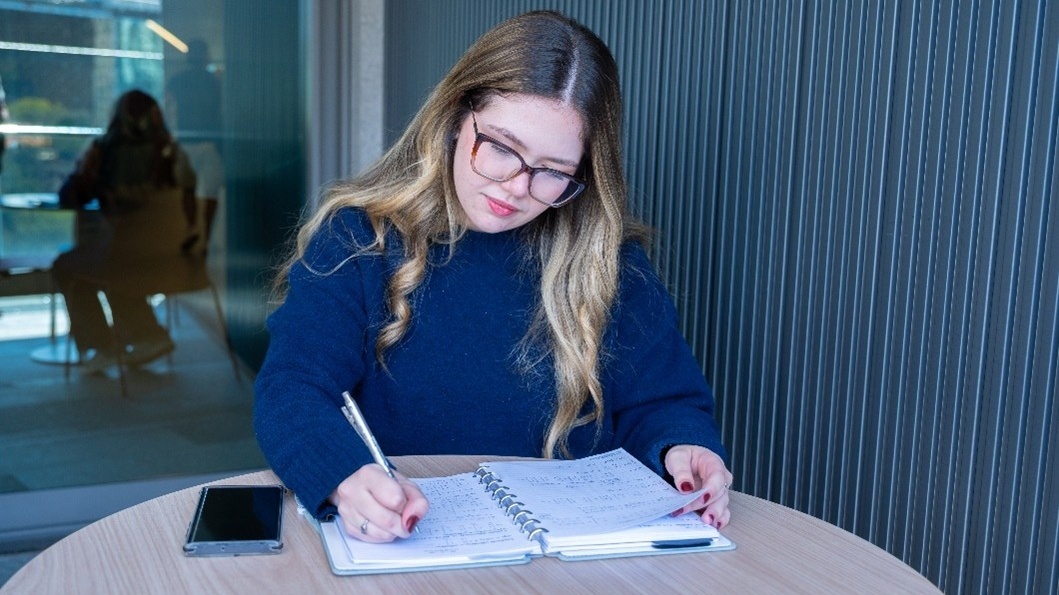 Estudante de óculos escreve em um caderno sobre uma mesa, em um ambiente de estudos.