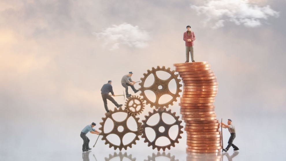 Miniaturas de trabalhadores ajustam engrenagens ao lado de uma pilha de moedas, com um homem no topo, simbolizando trabalho, economia e crescimento financeiro.
