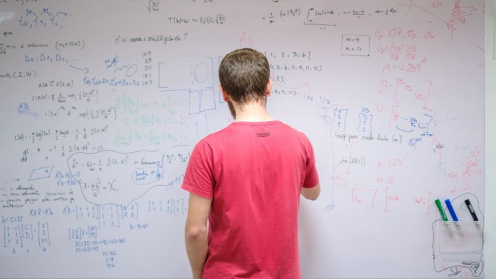 Pessoa em frente a um quadro branco com fórmulas, cálculos e anotações matemáticas em várias cores.
