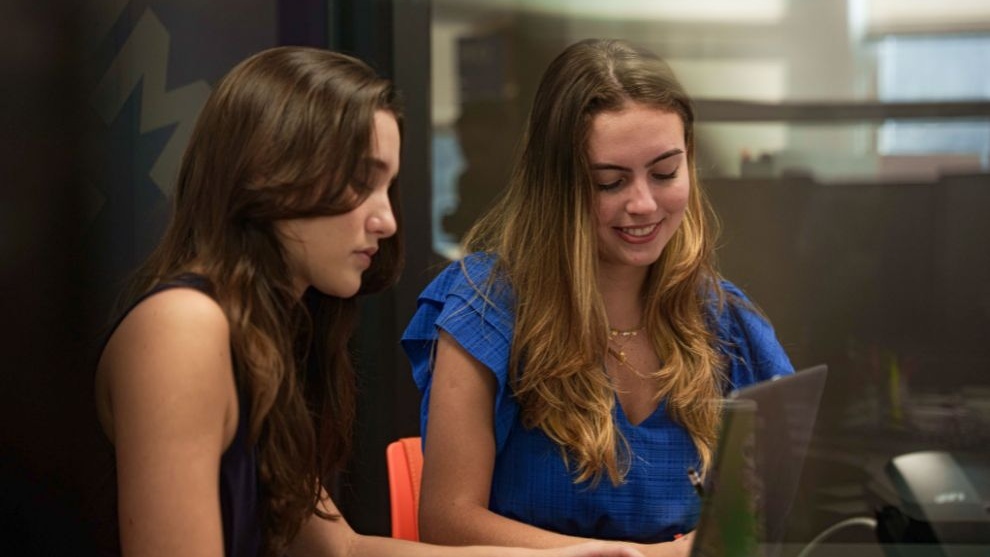 Duas jovens conversam em frente a um computador em um ambiente de estudo ou trabalho.