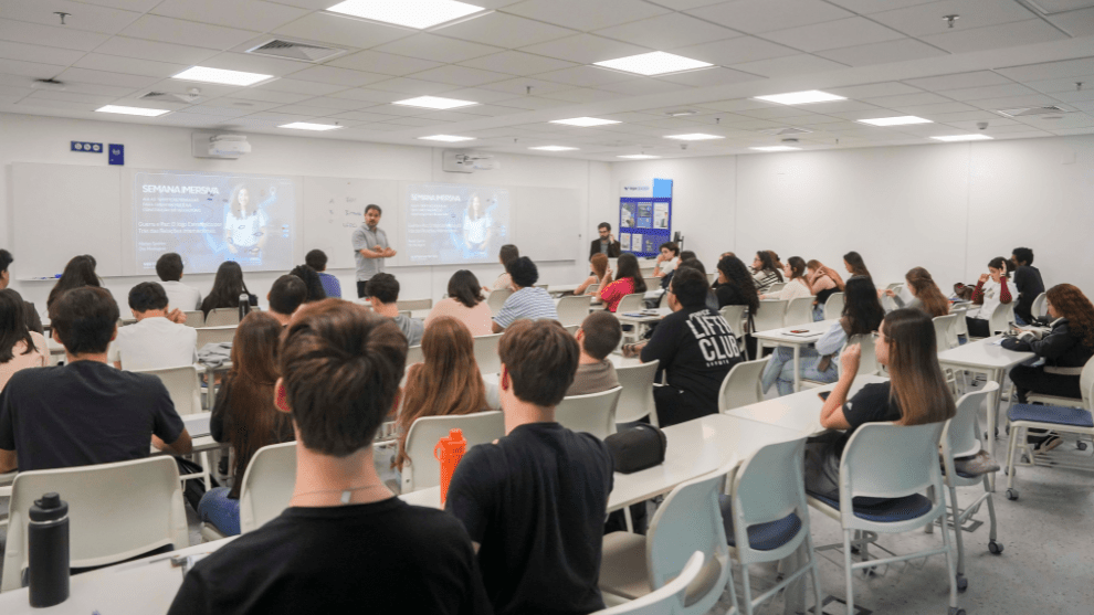 “Sala de aula universitária cheia de estudantes assistindo a uma apresentação conduzida por um professor à frente da turma.
