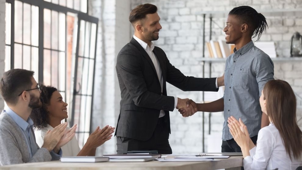 Um homem branco de terno e um homem negro de camisa casual trocam um aperto de mão caloroso em um escritório, sorrindo um para o outro. Outras pessoas sentadas ao redor em uma mesa aplaudem a interação.