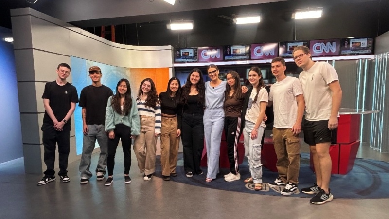 alunos em pé reunidos no canal de TV CNN ao lado de uma apresentadora de telejornal