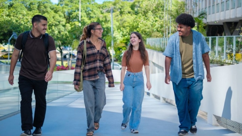 Quatro jovens caminham juntos ao ar livre em um campus, conversando e sorrindo, com árvores e prédios ao fundo.