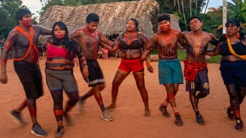 Um grupo de pessoas indígenas dançando em círculo em um terreiro de terra batida. Eles utilizam pinturas corporais tradicionais e trajes vibrantes, com uma maloca de palha e vegetação tropical ao fundo sob um céu claro.