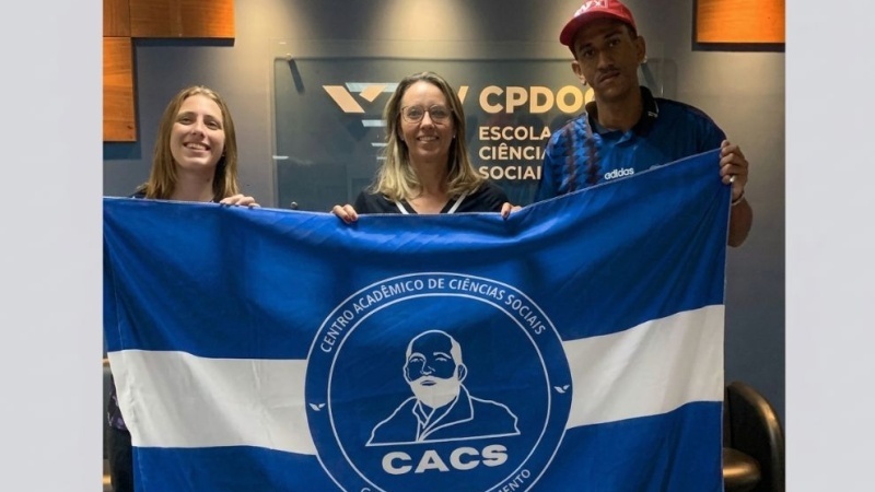 Três pessoas seguram uma bandeira azul e branca do Centro Acadêmico de Ciências Sociais Carlos Eduardo Sarmento (CACS)