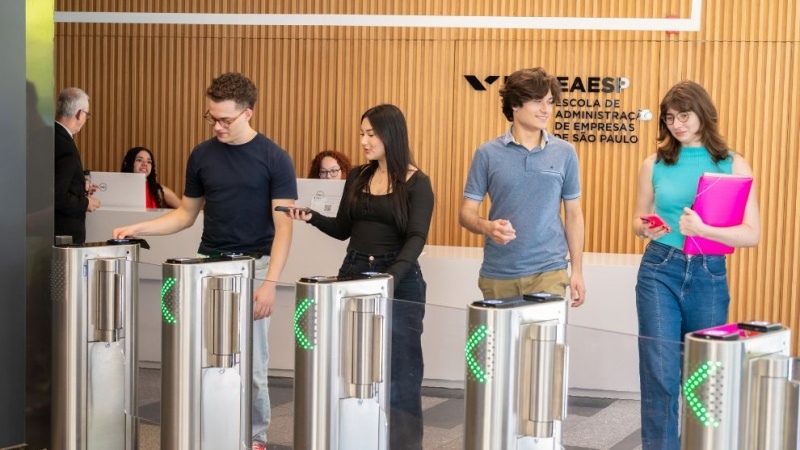 Quatro estudantes passando por catracas eletrônicas na entrada da FGV EAESP, segurando celulares e pastas, com recepção ao fundo