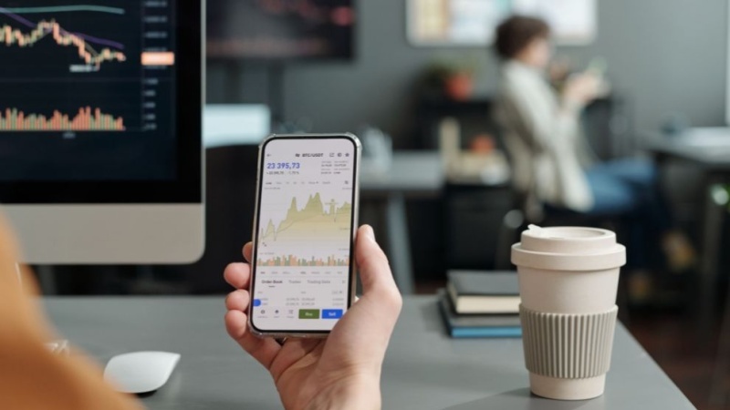 Pessoa acompanha gráficos de investimentos pelo celular, diante de um computador com indicadores financeiros.