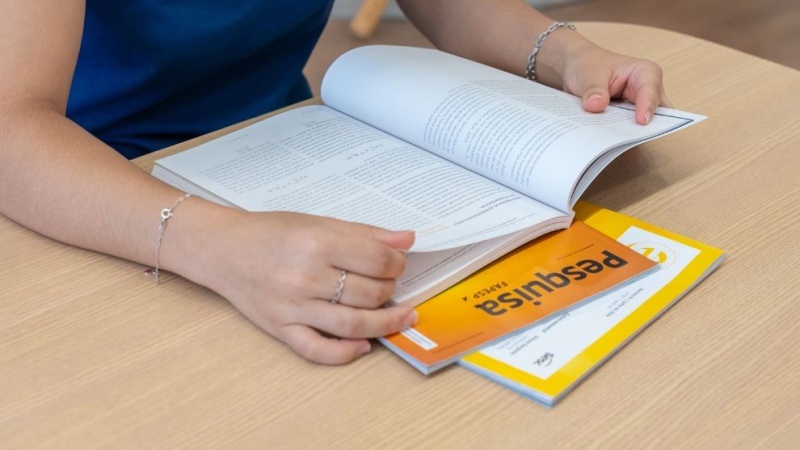 Pessoa lendo uma revista científica sobre a mesa, com foco nas mãos virando as páginas.