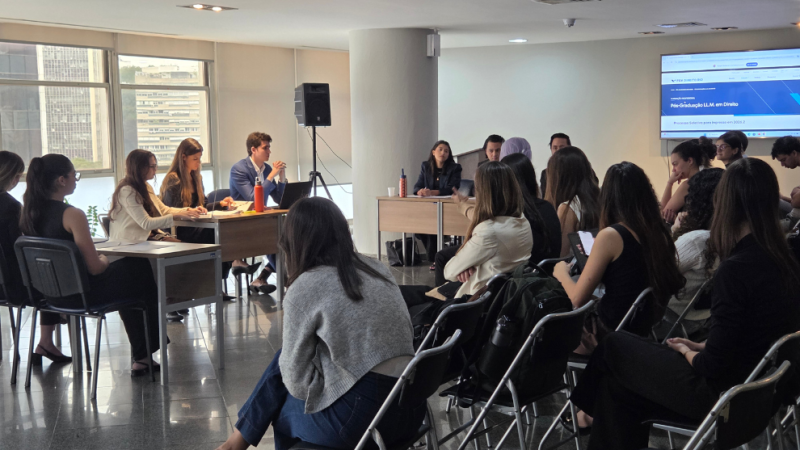 Grupo de estudantes participa de uma atividade acadêmica em sala, com apresentações em mesas e público sentado.
