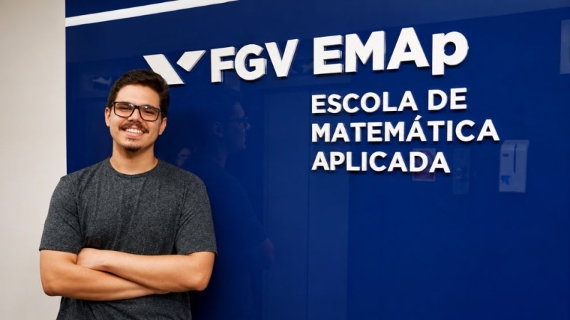 Rapaz sorrindo, de braços cruzados, em frente ao painel azul da FGV EMAP (Escola de Matemática Aplicada).