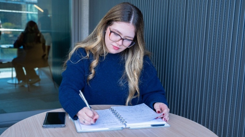Estudante de óculos escreve em um caderno sobre uma mesa, em um ambiente de estudos.