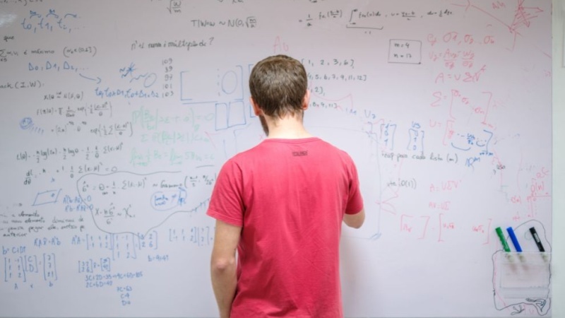 Pessoa em frente a um quadro branco com fórmulas, cálculos e anotações matemáticas em várias cores.