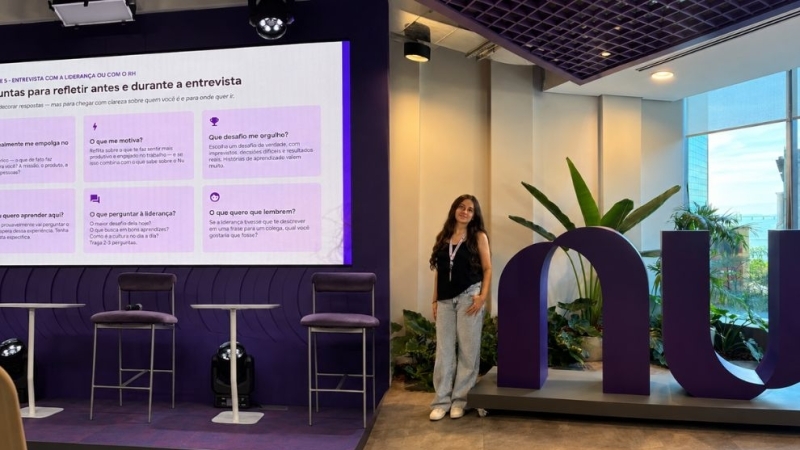 Ambiente moderno, com palco roxo, tela de apresentação, poltronas, plantas e grande logotipo “nu” da Nubank; uma aluna posa ao lado do logotipo.
