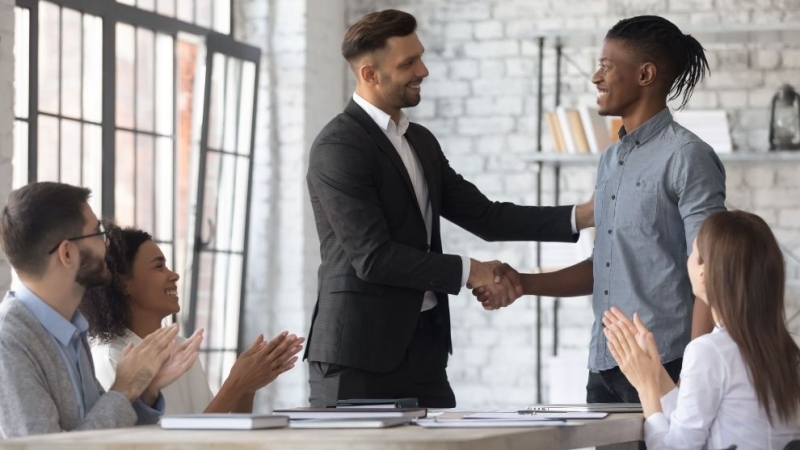 Um homem branco de terno e um homem negro de camisa casual trocam um aperto de mão caloroso em um escritório, sorrindo um para o outro. Outras pessoas sentadas ao redor em uma mesa aplaudem a interação.