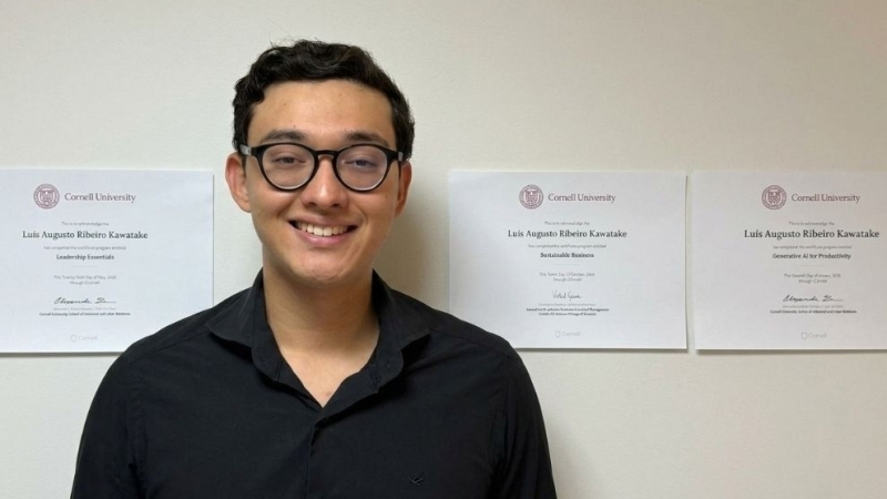 Um estudante sorrindo, vestindo camisa preta e óculos de armação escura, posando em formato horizontal com três certificados da Cornell University dispostos lado a lado ao fundo.