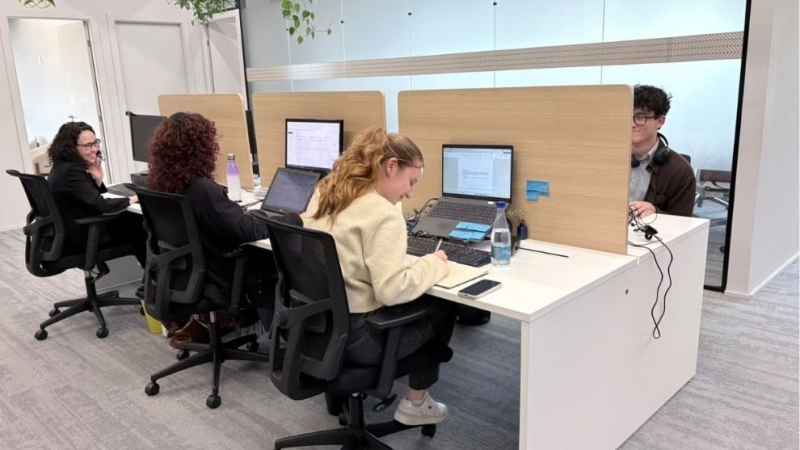 Escritório moderno e iluminado, com quatro pessoas trabalhando em mesas compartilhadas, usando notebooks e computadores. O ambiente possui divisórias de madeira, cadeiras de escritório, plantas suspensas e paredes de vidro.