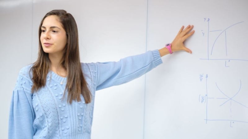 Uma mulher jovem de blusa azul-claro aponta para gráficos desenhados em um quadro branco ao seu lado.
