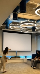 Professora apresenta conteúdo em uma sala de aula moderna enquanto estudantes acompanham a apresentação em um ambiente colaborativo.