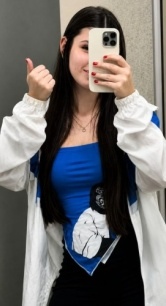 Jovem tirando uma selfie no espelho, usando jaqueta branca sobre uma blusa azul e fazendo sinal de positivo com o polegar.