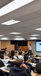 Sala de aula universitária com estudantes acompanhando uma apresentação de um professor em frente a uma tela de projeção, em um ambiente moderno e iluminado.