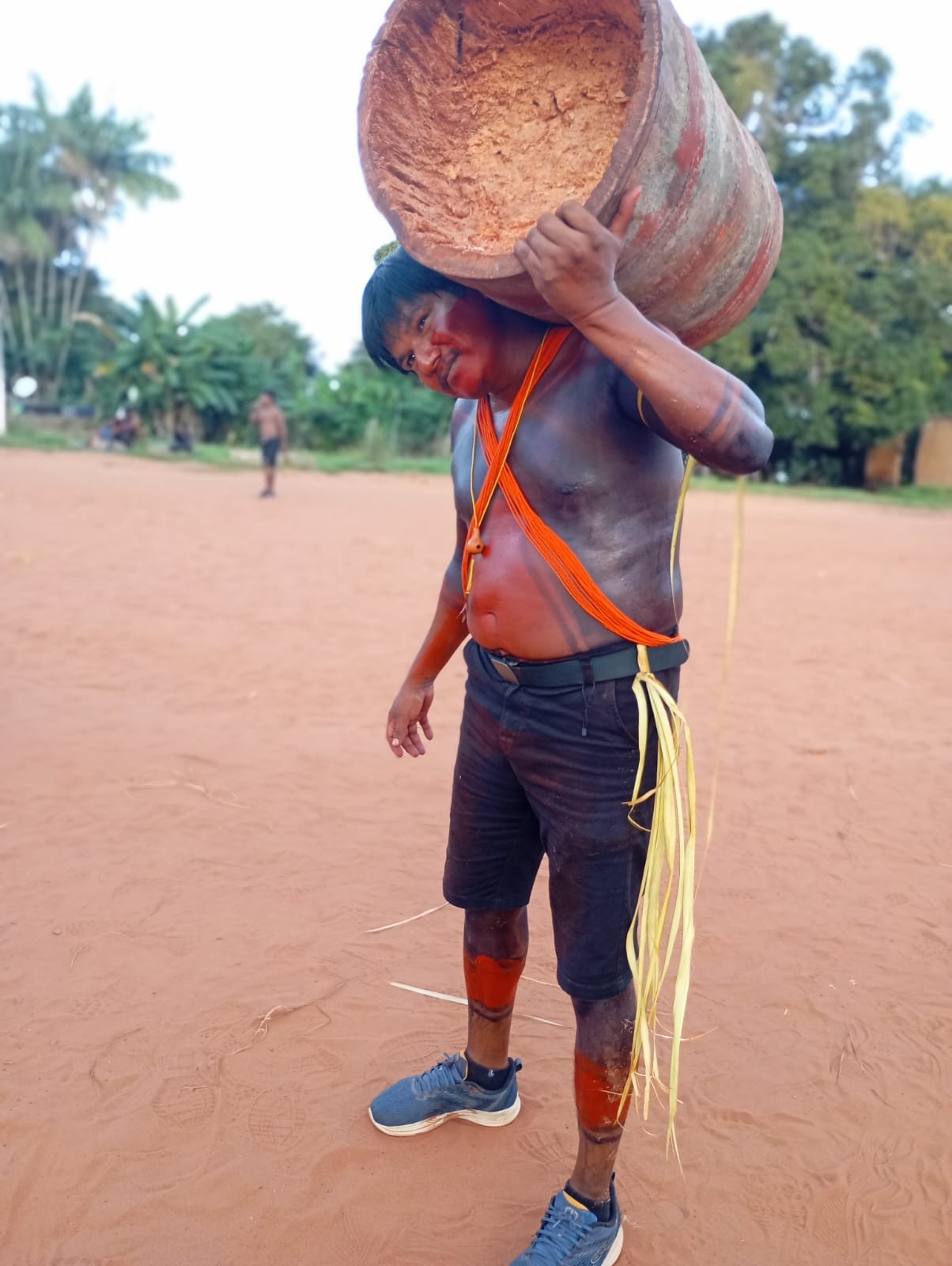 Homem indígena com pintura corporal vermelha carregando um grande tronco oco sobre o ombro em um pátio de terra batida.